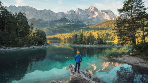 Ausblick auf das Börsenjahr 2026: Wo die Fäden zusammenlaufen Man enjoying the amazing morning scenery at a gorgeous lake in the Bavarian Alps, with teal water reflecting the view of the mountain range and the nice clouds
